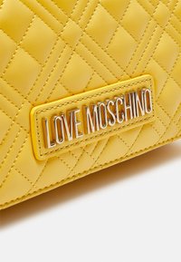 Žlutá prošívaná kabelka z umělé kůže se stříbrnou značkovou destičkou "LOVE MOSCHINO". Má šití ve tvaru diamantů a kontrastní žluté šití.