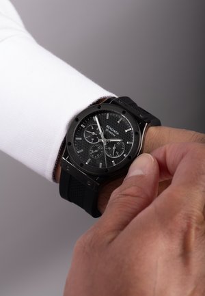 Schwarze analoge Uhr mit strukturiertem Zifferblatt, silbernen Akzenten und Gummiarmband, die an einem Handgelenk getragen wird. Die Uhr zeigt mehrere Unterzifferblätter und eine Datumsanzeige.