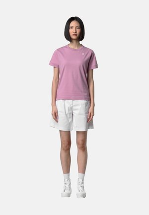 Giovane donna con capelli neri lisci che indossa una maglietta rosa a maniche corte, pantaloncini bianchi fino al ginocchio, calzini bianchi e scarpe da ginnastica bianche.