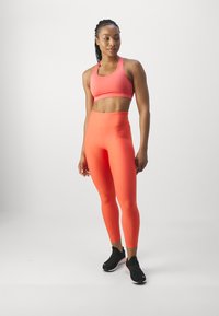 Korallfarget sportss Bras og matchende tights laget av elastisk, glatte stoff. Tightsene har høy midje og tettsittende design, kombinert med svarte joggesko.