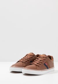 Brązowe skórzane trampki z białymi podeszwami, o fakturowanym wykończeniu, ze sznurówkami oraz czerwono-niebieskim paskiem na boku.