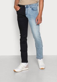 Two-tone jeans, left side black, right side light blue. Slim fit design, denim fabric, standard five-pocket style. White sneakers complete the look.