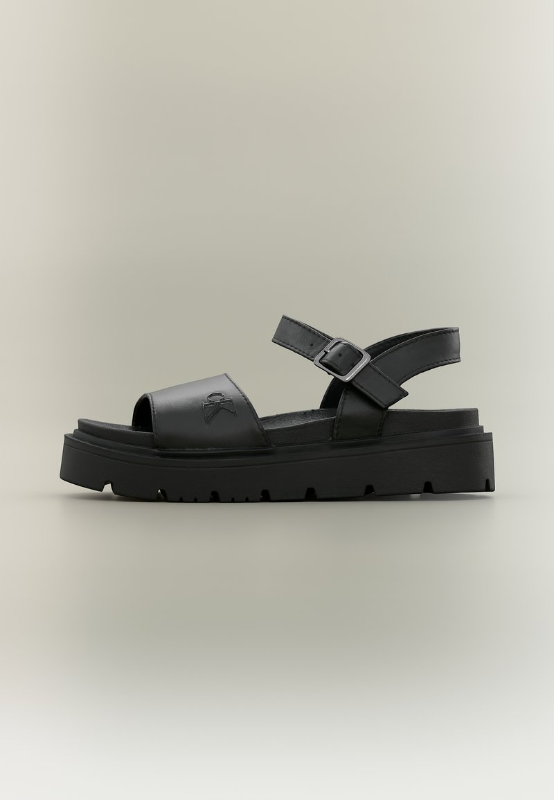 Μαύρο ανοιχτό σανδάλι με ρυθμιζόμενο λουράκι στον αστράγαλο, χοντρή σόλα και λογότυπο Calvin Klein ανάγλυφο στο μπροστινό λουράκι.