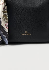 Black textured handbag with gold Anna Field logo and a floral patterned scarf draped on the left side against a white background.