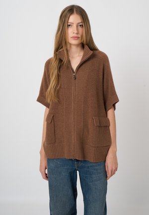Jonge vrouw met een bruine gebreide poncho met rits en voorzakken en blauwe spijkerbroek, staand tegen een effen lichte achtergrond.