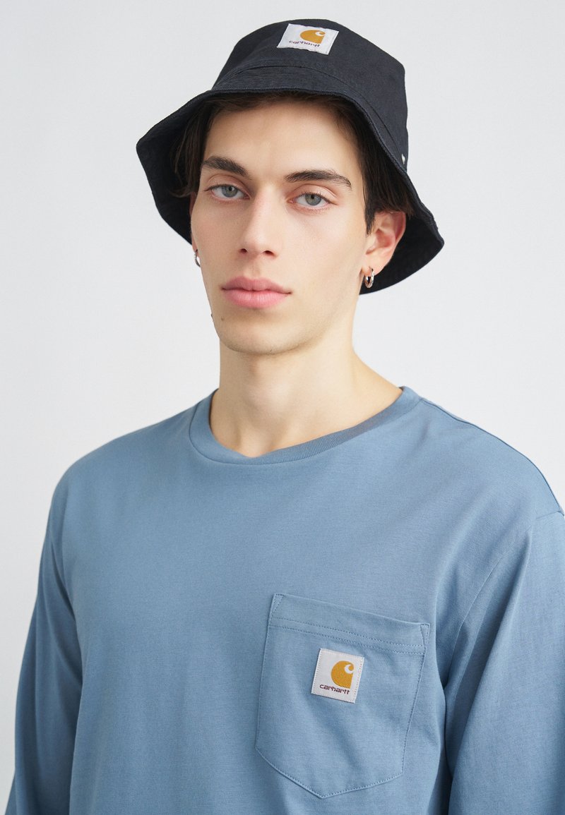 Jasnoniebieska koszula z długim rękawem z kieszonką na lewej piersi, na której znajduje się biała metka i pomarańczowe logo. Czarny kapelusz typu bucket z podobną metką.