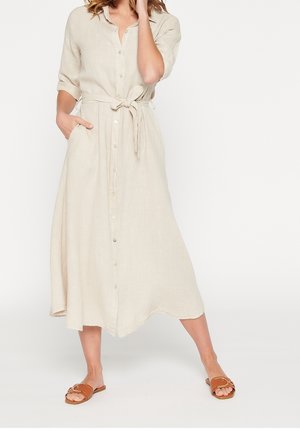 Kvinde iført beige knap-op midi kjole med taljebånd og opkørte ærmer, kombineret med brune flade sandaler.