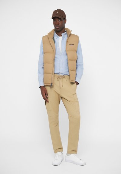 Gilet imbottito beige sopra una camicia azzurro chiaro a bottoni, abbinato a pantaloni cargo beige e scarpe da ginnastica bianche. Cappellino semplice marrone con dettaglio del logo.