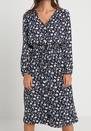 Kvinde iført en mørkeblå blomsterprint midi-kjole med lange ærmer, V-hals og en talje med bindebånd.