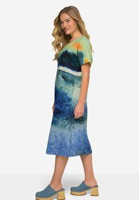 Grafisches Druckkleid mit blauen, grünen und orangenen Tönen, kurzen Ärmeln, wadenlanger Länge und glattem Stoffeindruck, kombiniert mit blauen Clogs.