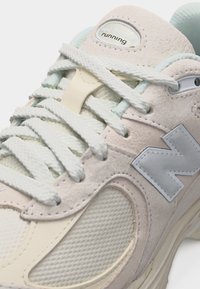 Chaussure de running New Balance beige et blanche avec des lacets texturés, des panneaux en suède et mesh, et un logo réfléchissant "N" sur le côté.