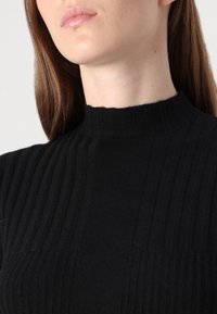 Femme aux longs cheveux bruns portant un pull noir à col roulé côtelé, montrant le cou et le bas du visage sur un fond uni.