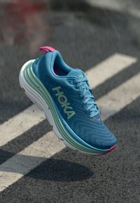 Bottine de sport bleue avec tige en maille texturée, marquage HOKA, accents roses et semelle blanche rembourrée avec des couches vertes.