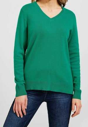 Femme portant un pull vert à col en V à manches longues, assorti à un jean bleu foncé, debout devant un fond uni clair.