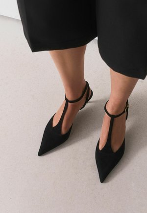Person trägt schwarze spitze T-Riemchen-Schuhe mit Knöchelriemen und steht auf hellgrauem Boden, gekleidet in schwarze, gekürzte Hosen.