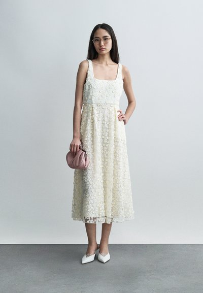 Μίντι φόρεμα από δαντέλα σε ελεφαντόδοντο με κεντήματα λουλουδιών, τετράγωνο ντεκολτέ και σιλουέτα γραμμής A. Συνδυασμένο με ροζ τσαντάκι χειρός και λευκά μυτερά φλατ παπούτσια.