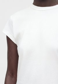 Personne portant un t-shirt blanc uni à manches courtes et col rond, visible du cou jusqu'au milieu du torse sur un fond gris clair.