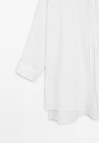 Blouse blanche à manches longues avec poignets boutonnés et un ourlet légèrement incurvé suspendue sur fond blanc.