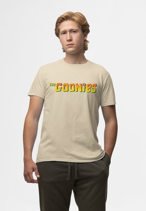 Jeune homme aux cheveux clairs portant un t-shirt beige « The Goonies » et un pantalon foncé, debout avec une main dans la poche devant un fond uni.