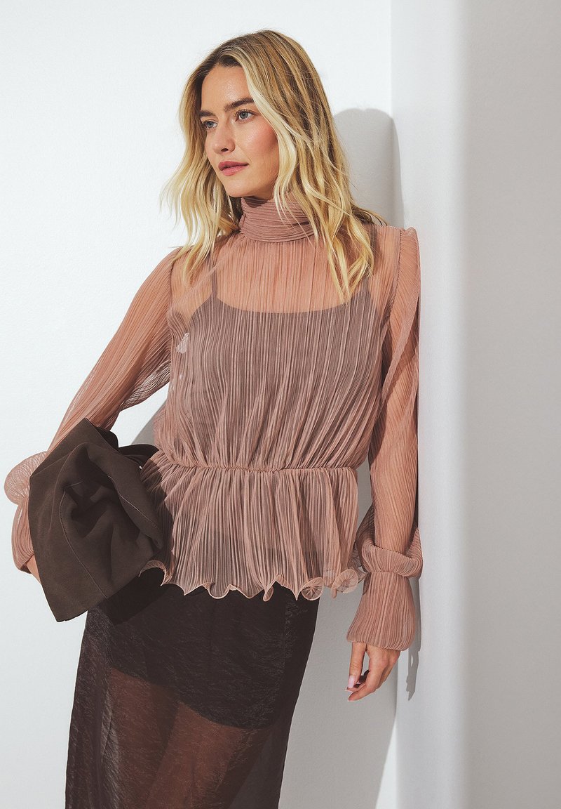 Caļš, plisēts tops maigos rozā toņos ar augstu apkakli, savilkta josta un plūstošām piedurknēm, savienojumā ar tumšu, caurspīdīgu svārku.