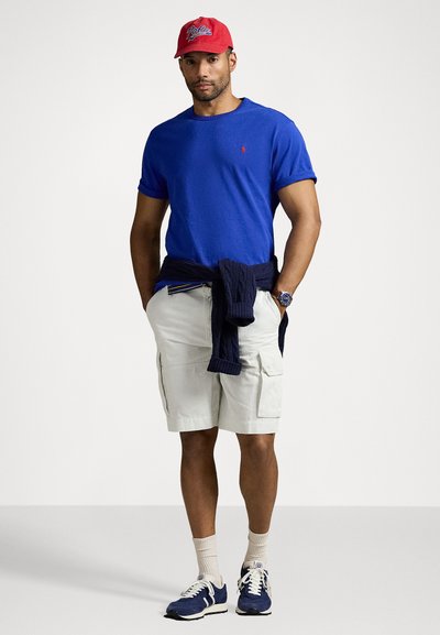 Μπλε βαμβακερό T-shirt με λογότυπο, μπεζ cargo σορτς, ναυτικό πουλόβερ δεμένο στη μέση, κόκκινο καπέλο μπέιζμπολ, ναυτικά αθλητικά παπούτσια με λευκές λεπτομέρειες.