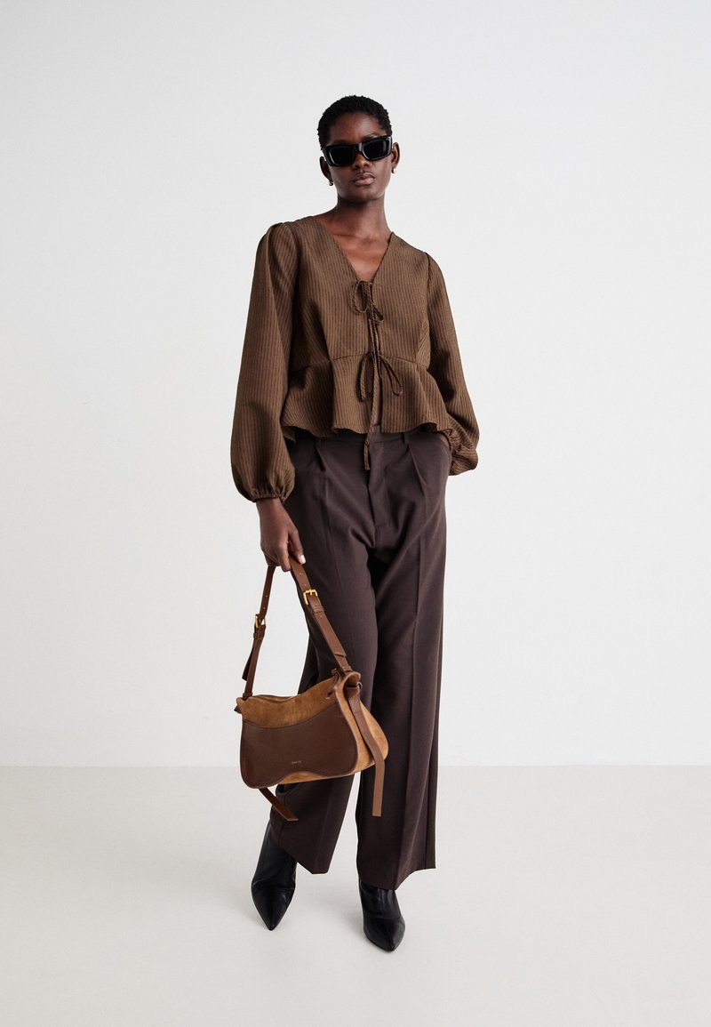 Brązowa bluzka o prążkowanej strukturze z wiązanym dekoltem, balonowymi rękawami, zestawiona z brązowymi spodniami o wysokim stanie oraz brązową skórzaną torebką. Czarne szpilki.
