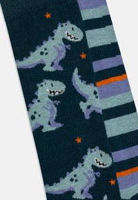 Barnesokker med en mørk teal-bunn som har dinosaurgrafikk og oransje stjerner på den ene siden, og et stripet mønster i blått og lilla på den andre siden.