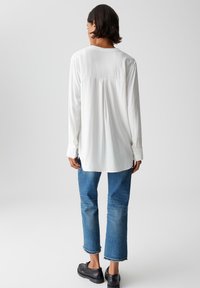 Persoon in een losse witte blouse met lange mouwen, blauwe cropped jeans met ruwe randen en zwarte loafers, staand met de achterkant naar de camera op een effen achtergrond.