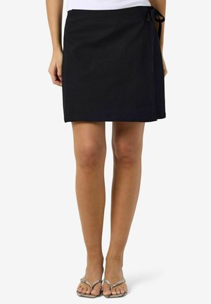 Kvinde iført et sort omslagsnederdel bundet i siden, kombineret med sølvfarvede sandaler med flettede stropper, stående foran en ensfarvet hvid baggrund.