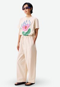 Mujer con pantalones anchos beige, camiseta beige con gran estampado de flor rosa, gafas de sol moradas y sandalias negras, de pie con una mano en el bolsillo.