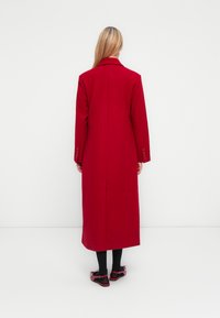 Long red coat with a tailored design, collar, and buttoned cuffs. Worn over black tights and red shoes with bows. Smooth texture.