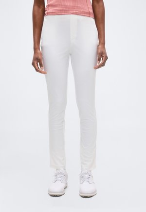 Osoba ubrana w dopasowane białe spodnie, białe sznurowane buty i różowy prążkowany top, stojąca na jednolitym jasnym tle.