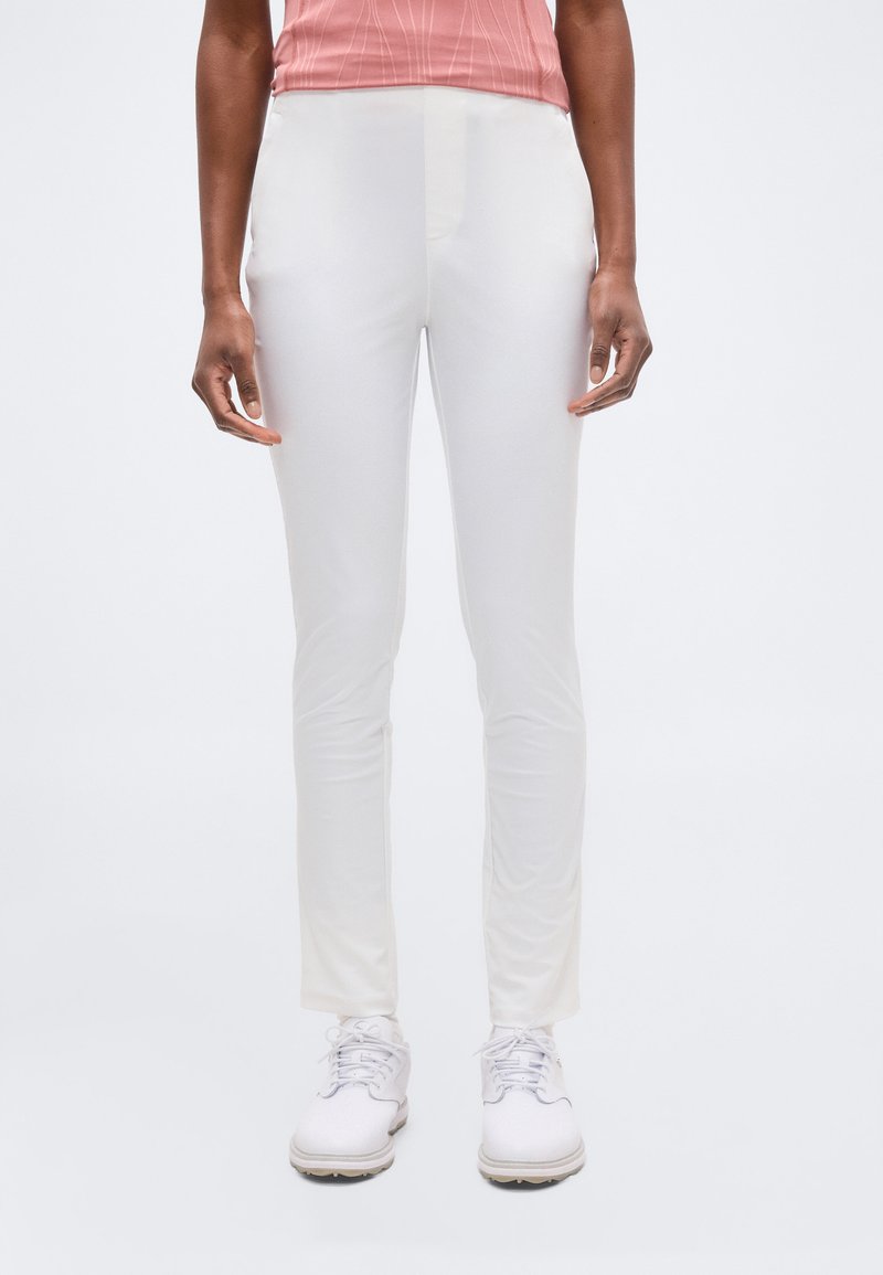 Άτομο που φοράει στενά λευκά παντελόνια, λευκά παπούτσια με κορδόνια και ροζ ριμπ μπλουζάκι, στέκεται μπροστά σε απλό φωτεινό φόντο.