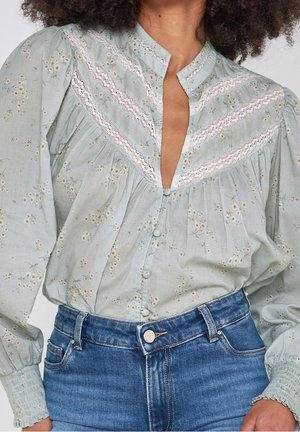 Femme portant un chemisier fleuri bleu clair avec des détails en dentelle et des manches bouffantes, rentrée dans un jean bleu taille haute.
