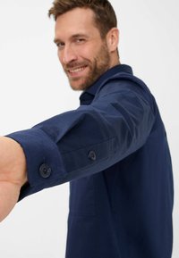 Chemise boutonnée bleu marine avec manches longues, présentant des poignets à boutons et une poche à l'avant. Tissu en coton texturé.