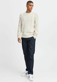 Suéter de punto trenzado en color crema con cuello y puños acanalados, combinado con pantalones de mezclilla azul oscuro y zapatillas blancas con rayas negras.
