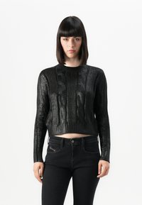 Femme avec des cheveux raides et foncés portant un pull en maille torsadée noir à manches courtes et un jean noir taille haute, se tenant devant un fond blanc uni.
