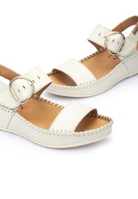 Sandales en cuir blanc avec bout ouvert, sangle à boucle et couture décorative le long de la semelle. Présentent un coussin intégré pour le confort.