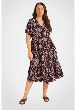 Kvinde iført en sort midi-kjole med lyserønt blomsterprint, korte ærmer, knapper foran og brune sandaler, stående mod en ensfarvet baggrund.