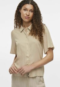 Beige kortærmet bluse med krave, blødt stof, knaplukning og snøre detaljer ved kanten. Glat tekstur.