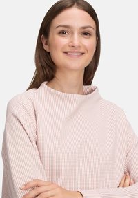Junge Frau mit langen braunen Haaren, die lächelt und ein strukturiertes, hellrosa und weiß kariertes Langarmshirt trägt, mit verschränkten Armen.