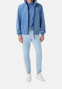 Veste zippée bleu clair sur un polo assorti, accompagnée de pantalons roulés bleu clair et de baskets blanches. Tissu lisse et coupe ajustée.