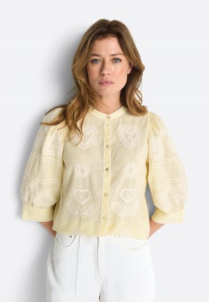 Žena so svetlohnedými vlasmi má na sebe svetložltú vyšívanú blúzku s nafúknutými rukávmi a biele nohavice, stojí na svetlom pozadí.