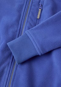 Sininen fleece-takki, jossa on teksturoitu pinta, täysi vetoketju ja ribbetty hihansuut. Huomiota herättäviä yksityiskohtia ovat sileä vetoketjunauha.