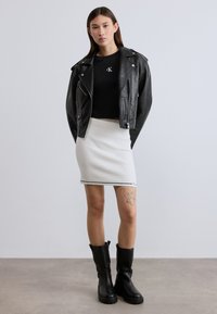 Μαύρο δερμάτινο μπουφάν μοτοσικλετιστή πάνω από μαύρο μπλουζάκι με λογότυπο, συνδυασμένο με λευκή ριμπ λευκή μίνι φούστα και μαύρες μπότες μέχρι το γόνατο.