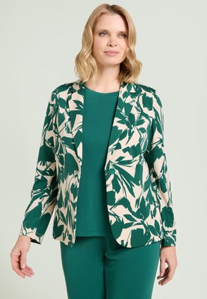 Vrouw draagt groene broek en top met crème blazer met donker groene abstracte bladpatronen, staand tegen een bleke achtergrond.