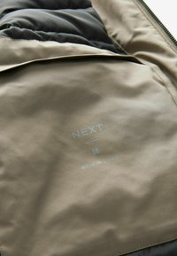 O țesătură khaki, cu o textură netedă, prezintă un eticheta care spune "NEXT M MADE IN CHINA", plasată pe un căptușal mai închis la culoare.