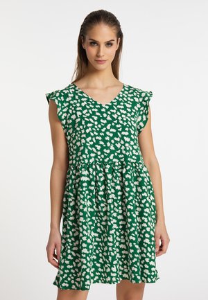 Vrouw draagt een groene mouwloze jurk met wit abstract bloemenpatroon en een V-hals, staand tegen een effen witte achtergrond.