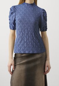 Halenka s krátkým rukávem vyrobená z texturované modré látky s diamantovým vzorem, s vysokým výstřihem a nabíranými rukávy, v kombinaci s hnědou sukní.