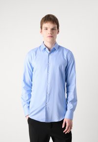 Camisa de botón azul claro hecha de una tela suave, con un collar clásico y mangas largas. El modelo está de pie contra un fondo simple.